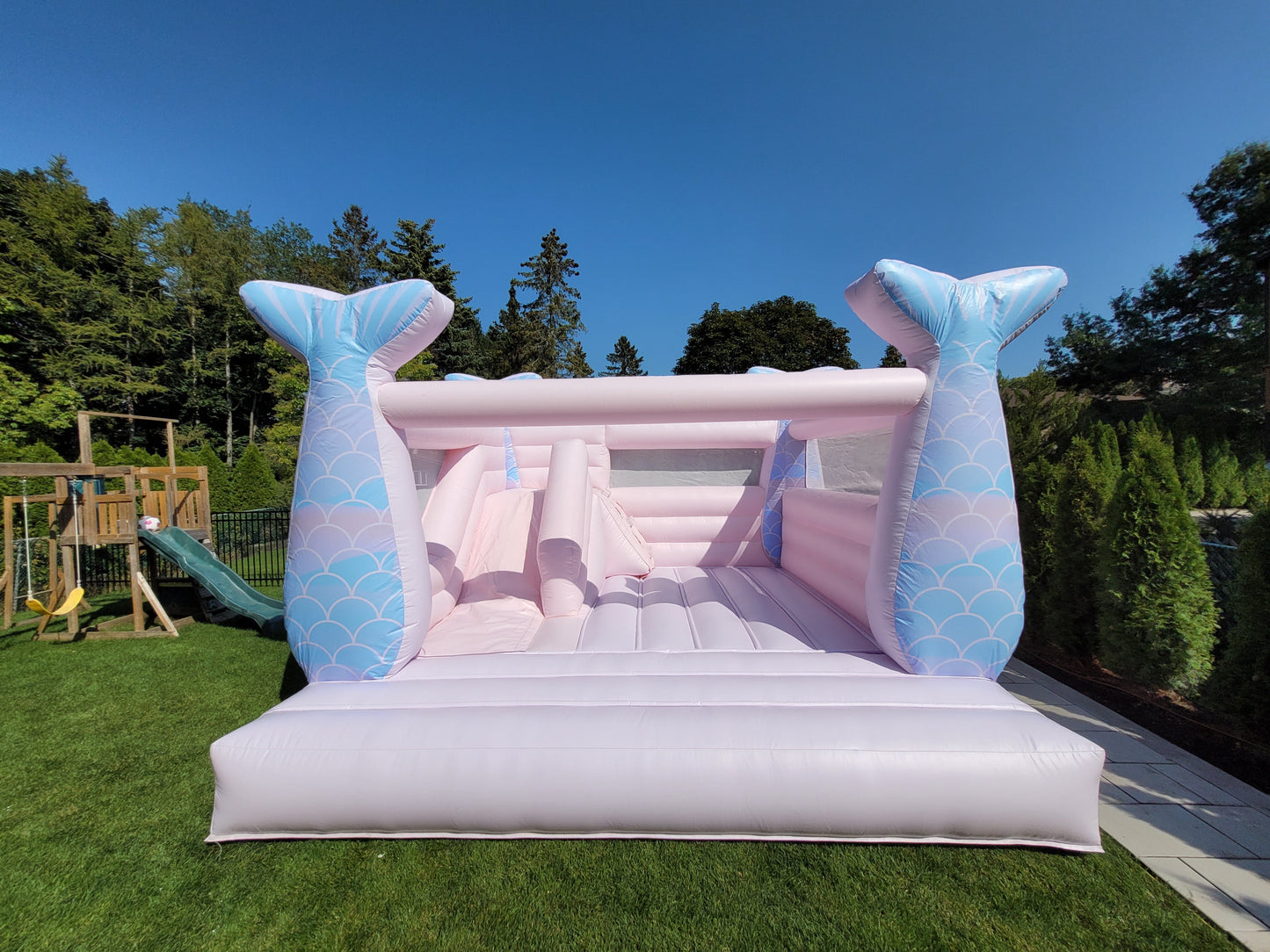 Inflatable bounce house with mermaid tail design on a grassy area with trees in the background